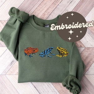 May include: A green sweatshirt with three embroidered frogs in orange, blue, and yellow. The sweatshirt has the word "Embroidered" in a brown circle with stars.