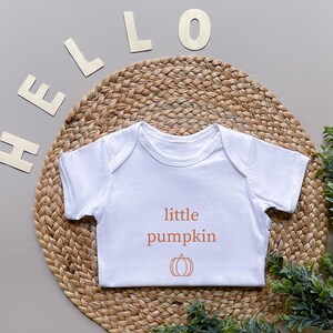 May include: A white baby onesie with the words "little pumpkin" and a pumpkin graphic in orange. The onesie is on a woven mat with the word "HELLO" spelled out in wooden letters.