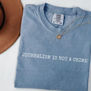 May include: A blue t-shirt with the text "JOURNALISM IS NOT A CRIME" in white. The shirt has a crew neck and short sleeves. A gold necklace with a pendant is draped across the shirt. A brown hat is visible in the upper left corner.