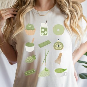 以下が含まれることがあります： 抹茶をテーマにした緑色のデザインが施された白いTシャツ。デザインには、アイスクリーム、ケーキ、ドリンクなど、さまざまな抹茶のおやつが描かれています。