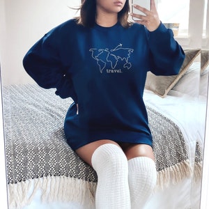 Puede incluir: Sudadera azul marino con un contorno blanco de un mapa del mundo y un avi&oacute;n. La palabra "travel" est&aacute; impresa debajo del mapa. La sudadera es de manga larga y tiene cuello redondo.
