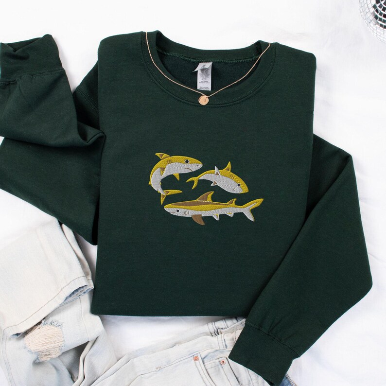 Pode incluir: Uma camisola verde escura com um design bordado amarelo de tr&ecirc;s tubar&otilde;es. Os tubar&otilde;es est&atilde;o a nadar em grupo, com as suas barbatanas e caudas vis&iacute;veis.