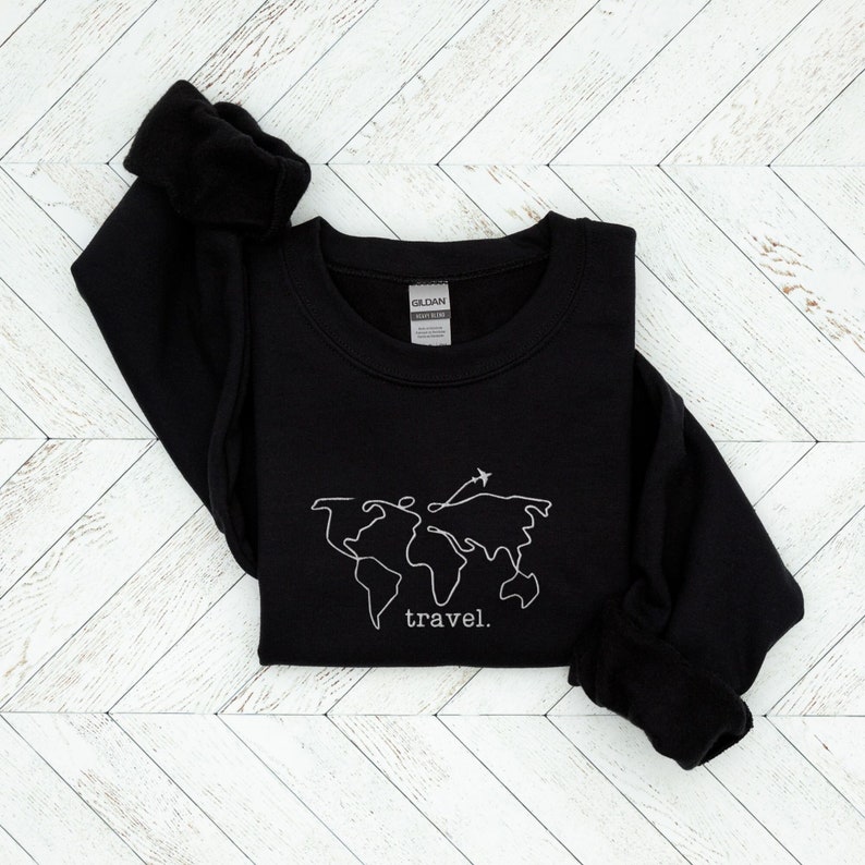 Puede incluir: Sudadera negra con un contorno blanco de un mapa del mundo y un avi&oacute;n. La palabra "travel" est&aacute; escrita debajo. La sudadera est&aacute; hecha de un material suave y tiene cuello redondo.