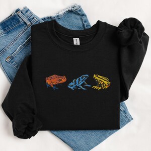 May include: Black sweatshirt with a graphic of three colorful frogs. The frogs are in a line, facing the right. The first frog is orange, the second is blue and black, and the third is yellow.