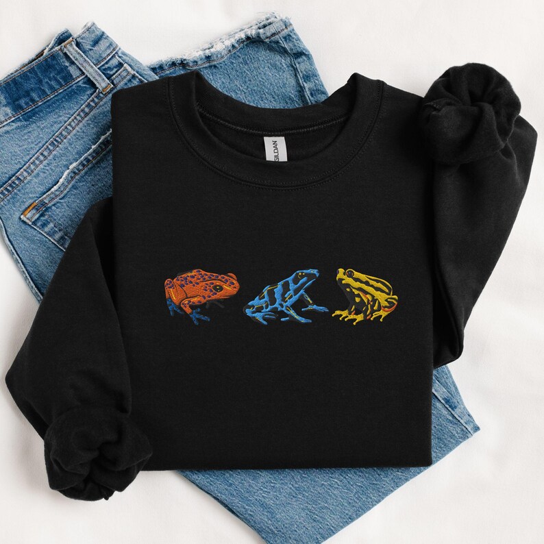 May include: Black sweatshirt with a graphic of three colorful frogs. The frogs are in a line, facing the right. The first frog is orange, the second is blue and black, and the third is yellow.