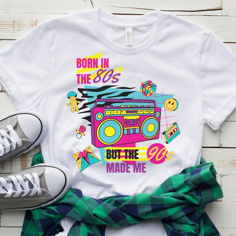 May include: White t-shirt with a graphic design featuring a boombox, a Rubik's cube, a pacifier, a baby doll, a cassette tape, and a smiley face. The text on the shirt reads "Born in the 80s But the 90s Made Me".