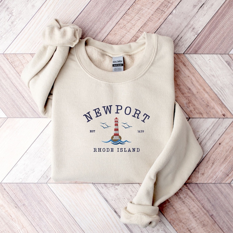May include: A beige sweatshirt with a navy blue graphic of a lighthouse, seagulls, and the text "NEWPORT EST 1639 RHODE ISLAND".