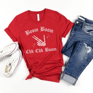 May include: Red t-shirt with a white skeleton hand making a peace sign and the text "Boom Boom Chk Chk Boom" in a gothic font.