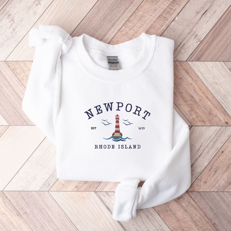May include: White sweatshirt with a blue and red lighthouse graphic and the text "NEWPORT EST 1639 RHODE ISLAND".