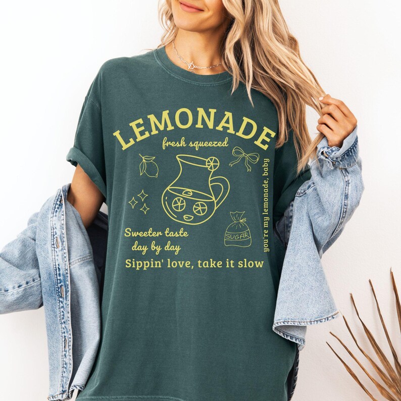 Puede incluir: Camiseta verde oscuro con la palabra "LEMONADE" en amarillo, junto con ilustraciones de limones, una jarra y una bolsa de az&uacute;car. La camiseta tambi&eacute;n presenta las frases "fresh squeezed", "Sweeter taste day by day" y "Sippin' love, take it slow".