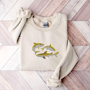 Pode incluir: Uma camisola de cor creme com um desenho bordado de tr&ecirc;s tubar&otilde;es amarelos. Os tubar&otilde;es est&atilde;o a nadar em dire&ccedil;&otilde;es diferentes. A camisola tem um decote redondo e mangas compridas.