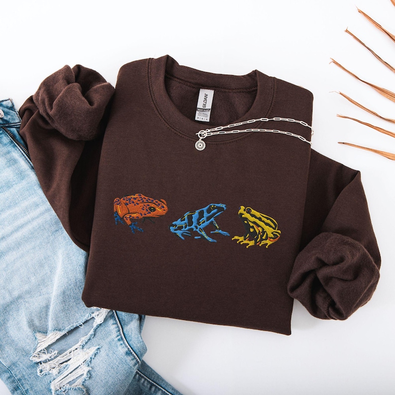 May include: A brown sweatshirt with a graphic of three frogs in different colors. The frogs are in a line, facing the right. The sweatshirt has a crew neck and long sleeves.