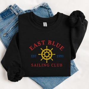 May include: Black sweatshirt with the text "EAST BLUE SAILING CLUB" in red and blue, with a yellow ship's wheel graphic. The sweatshirt also has the text "EST 1999". The sweatshirt is folded on top of a pair of blue jeans.