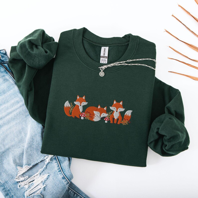 May include: A dark green sweatshirt with a crew neck and embroidered foxes. The foxes are orange and have white accents. The sweatshirt has a silver chain necklace with a pendant.