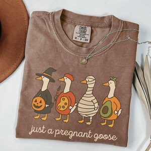 Può includere: T-shirt marrone Comfort Colors con un design di Halloween di quattro oche in costume: una strega, un artista, una mummia e un avocado. Il testo "just a pregnant goose" è stampato sotto.