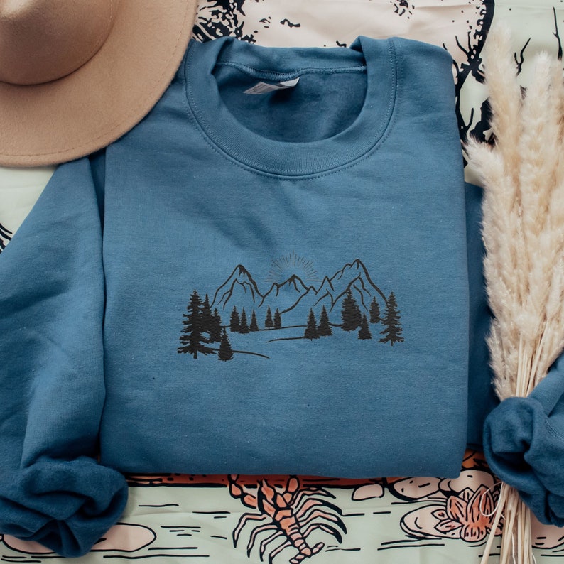 Op de afbeelding: Een blauw sweatshirt met een zwarte grafische afbeelding van een bergketen en bomen. De afbeelding is gecentreerd op de voorkant van het sweatshirt.