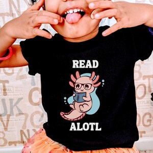 May include: A black t-shirt with a white axolotl wearing glasses and reading a book. The text "READ ALOTL" is printed on the shirt.