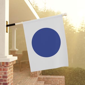 May include: A white flag with a large, solid blue circle in the center. The flag is hanging from a pole outside a house.