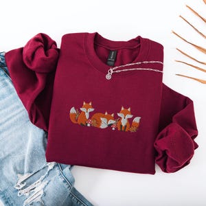 May include: A maroon sweatshirt with a crew neck and long sleeves features an embroidered design of three foxes with orange fur and white accents. The foxes are surrounded by small green leaves and red mushrooms.