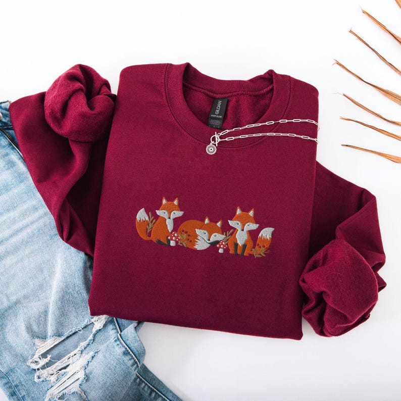 May include: A maroon sweatshirt with a crew neck and long sleeves features an embroidered design of three foxes with orange fur and white accents. The foxes are surrounded by small green leaves and red mushrooms.
