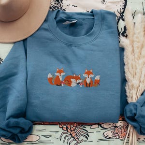 May include: A blue sweatshirt with an embroidered design of three foxes with red accents. The foxes are surrounded by small red mushrooms.