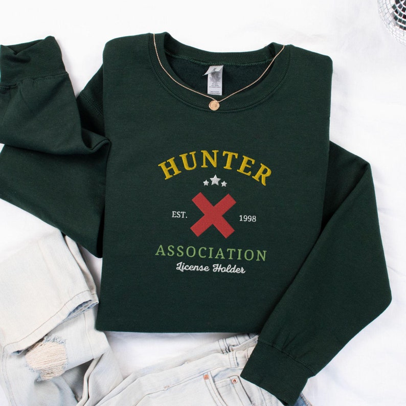 May include: A dark green sweatshirt with the text "HUNTER ASSOCIATION License Holder" and a red X with three yellow stars above it. The text "EST. 1998" is below the X.
