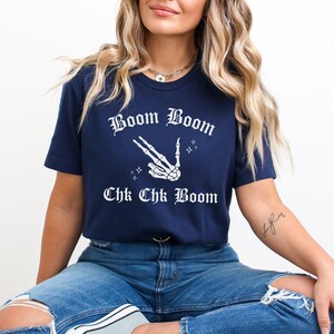 May include: A navy blue t-shirt with a white skeleton hand making a peace sign and the text "Boom Boom Chk Chk Boom" in a gothic font.