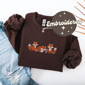 May include: A brown sweatshirt with an embroidered design of three foxes. The foxes are brown with orange accents and are surrounded by green leaves and mushrooms. The sweatshirt has a crew neck and long sleeves. The text "Embroidered" is written in a white circle with a brown background.