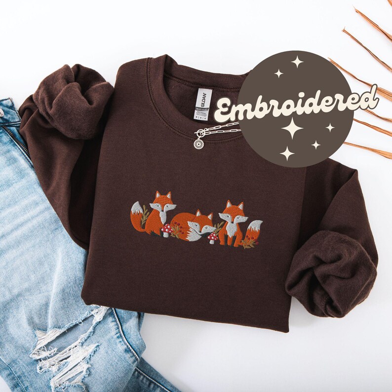 May include: A brown sweatshirt with an embroidered design of three foxes. The foxes are brown with orange accents and are surrounded by green leaves and mushrooms. The sweatshirt has a crew neck and long sleeves. The text "Embroidered" is written in a white circle with a brown background.