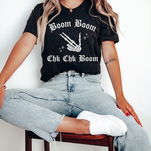 May include: A dark grey t-shirt with a white skeleton hand making a peace sign and the text "Boom Boom Chk Chk Boom" in a gothic font.