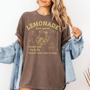 Puede incluir: Una camiseta marr&oacute;n con la palabra "LEMONADE" en amarillo, junto con un gr&aacute;fico de una jarra, limones y el texto "fresh squeezed". El dise&ntilde;o tambi&eacute;n incluye "Sweeter taste day by day" y "Sippin' love, take it slow". La camiseta se lleva con una chaqueta vaquera.