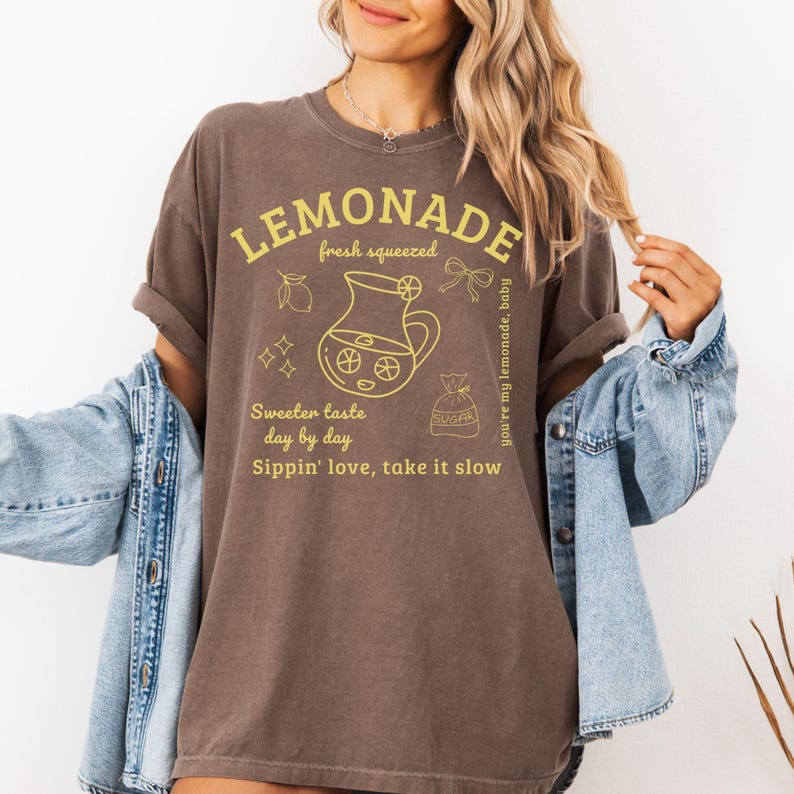 Puede incluir: Una camiseta marr&oacute;n con la palabra "LEMONADE" en amarillo, junto con un gr&aacute;fico de una jarra, limones y el texto "fresh squeezed". El dise&ntilde;o tambi&eacute;n incluye "Sweeter taste day by day" y "Sippin' love, take it slow". La camiseta se lleva con una chaqueta vaquera.