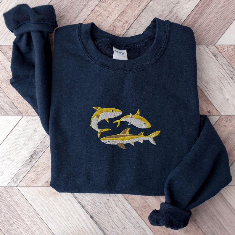Pode incluir: Uma camisola azul marinho com um design bordado de tr&ecirc;s tubar&otilde;es amarelos. Os tubar&otilde;es est&atilde;o a nadar em grupo, com as caudas viradas para o observador.