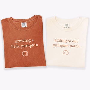 May include: Two folded t-shirts, one rust-colored and one cream-colored, with the text "growing a little pumpkin" and "adding to our pumpkin patch" printed on them. Each shirt has a pumpkin graphic below the text. The shirts are likely for a pregnancy announcement or baby shower.