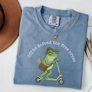 May include: A light blue Comfort Colors t-shirt with the phrase "Off to defend the free press" encircling a cartoon frog riding a scooter. The frog wears glasses and a hat, carrying a bag and newspaper. A gold necklace is draped across the shirt.