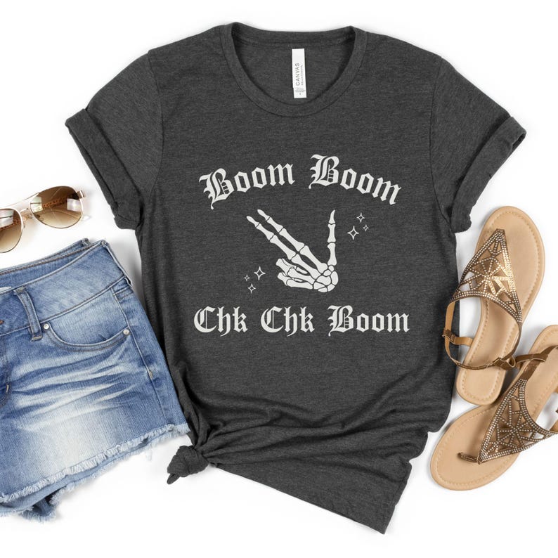 May include: A dark gray t-shirt with a white graphic print of a skeleton hand making a peace sign with the text "Boom Boom Chk Chk Boom" in a gothic font.