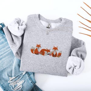 May include: A gray sweatshirt with an embroidered design of three foxes. The foxes are orange and brown with white accents. The foxes are arranged in a line, with the first fox looking to the right, the second fox looking to the left, and the third fox looking to the right. The sweatshirt is on a white surface.