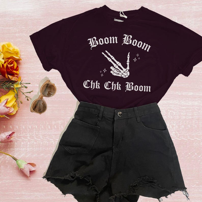 May include: A black t-shirt with white text that reads 'Boom Boom Chk Chk Boom' and a white skeleton hand making a peace sign. The t-shirt is paired with black denim shorts.