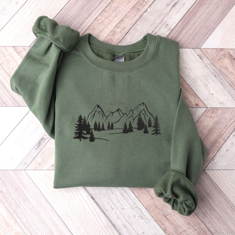 Op de afbeelding: Een groen sweatshirt met een zwarte berg- en boomprint. De afbeelding toont een bergketen met een opkomende zon erachter.