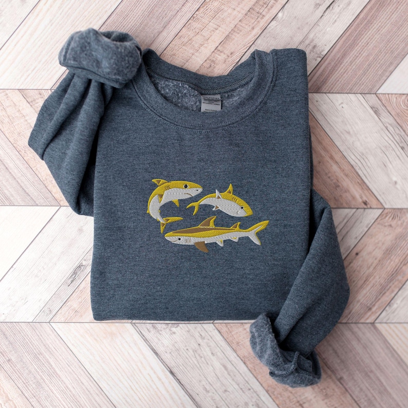 Pode incluir: Uma camisa de moletom cinza escuro com um design bordado de tr&ecirc;s tubar&otilde;es amarelos e brancos.