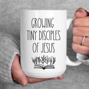 May include: White ceramic mug with the text "GROWING TINY DISCIPLES OF JESUS" in black, along with a graphic of an open book with plants. The mug is held by a person wearing a gray sweater.