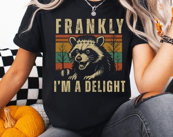 May include: Black t-shirt with a raccoon graphic and the text "FRANKLY I'M A DELIGHT" in a retro style. The raccoon is set against a sunset-colored background with trees. A casual, comfortable tee.