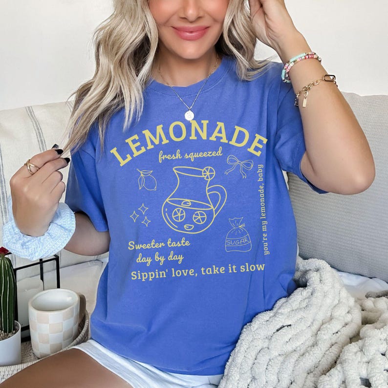 Puede incluir: Una camiseta azul con un gr&aacute;fico amarillo que dice "LEMONADE" y "fresh squeezed". El gr&aacute;fico incluye una jarra, rodajas de lim&oacute;n y las frases "Sweeter taste day by day" y "Sippin' love, take it slow."