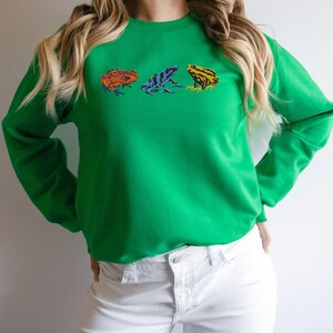 May include: A green sweatshirt with a row of three colorful frog illustrations printed on the front.