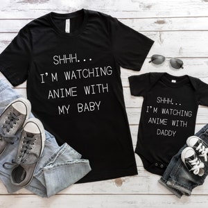 May include: A black t-shirt with the text "SHHH... I'M WATCHING ANIME WITH MY BABY" in white. A black baby onesie with the text "SHHH... I'M WATCHING ANIME WITH DADDY" in white. Both shirts are on a white wooden background with blue jeans and gray sneakers.