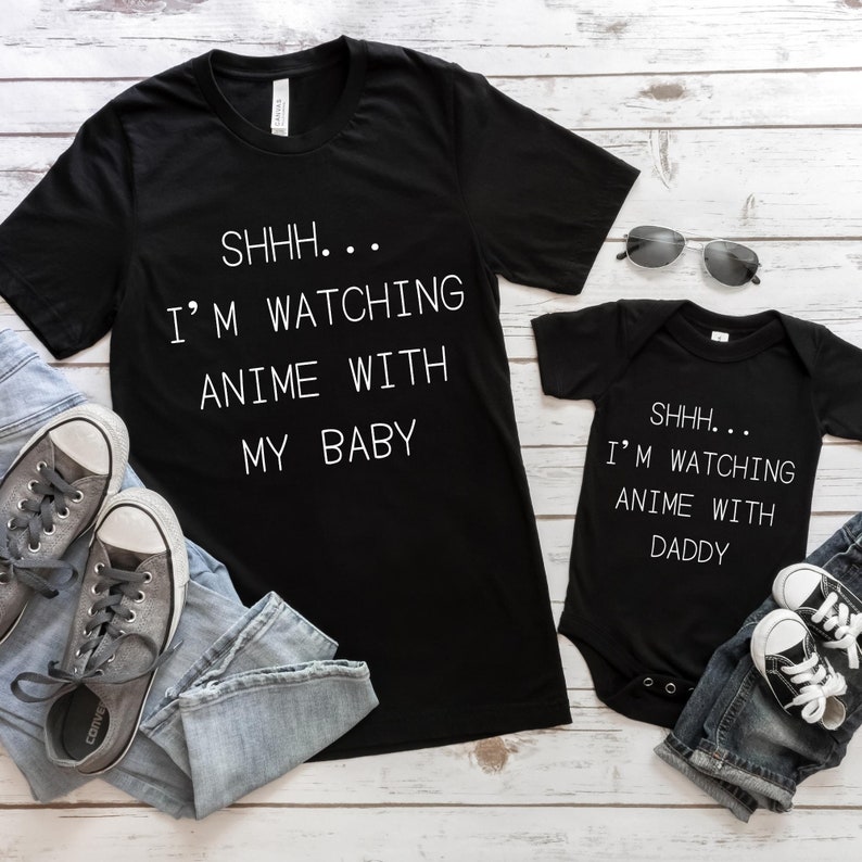 May include: A black t-shirt with the text "SHHH... I'M WATCHING ANIME WITH MY BABY" in white. A black baby onesie with the text "SHHH... I'M WATCHING ANIME WITH DADDY" in white. Both shirts are on a white wooden background with blue jeans and gray sneakers.