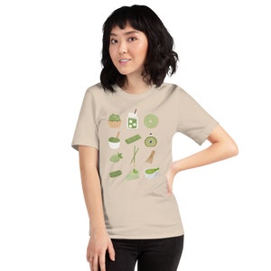 以下が含まれることがあります： 抹茶をテーマにした様々なアイテムが描かれた、ベージュのTシャツ。抹茶のカップ、抹茶アイス、抹茶ケーキ、抹茶ロールなどが描かれている。