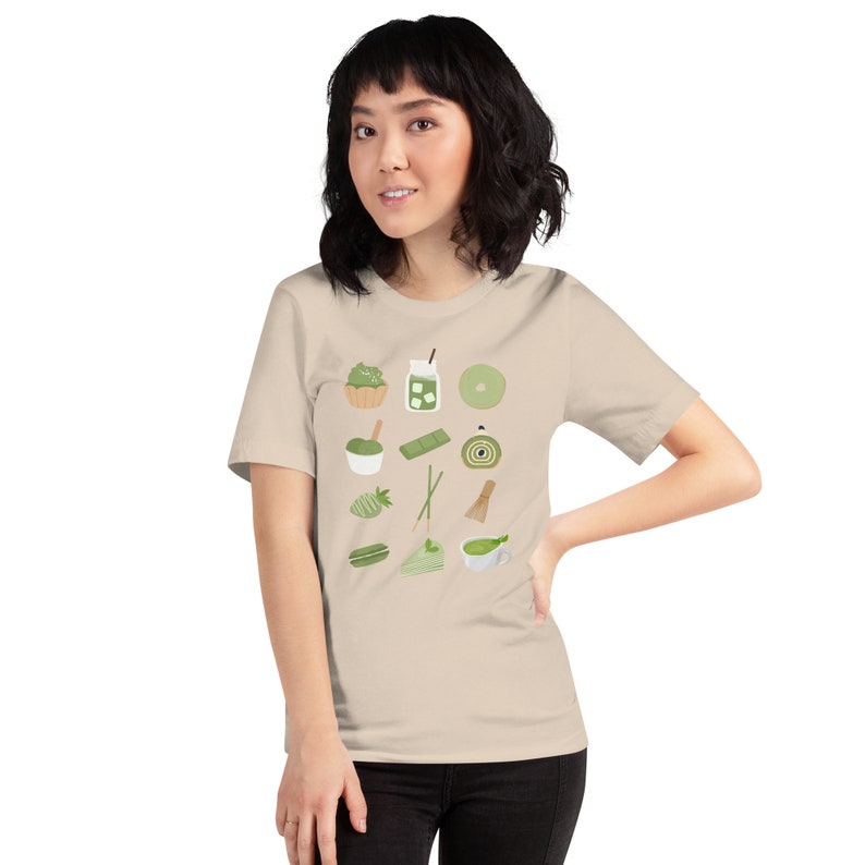 以下が含まれることがあります： 抹茶をテーマにした様々なアイテムが描かれた、ベージュのTシャツ。抹茶のカップ、抹茶アイス、抹茶ケーキ、抹茶ロールなどが描かれている。
