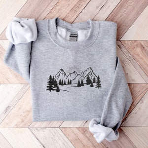 Op de afbeelding: Een grijs sweatshirt met een zwarte print van een bergketen en bomen. De afbeelding toont een zon die opkomt achter de bergen.