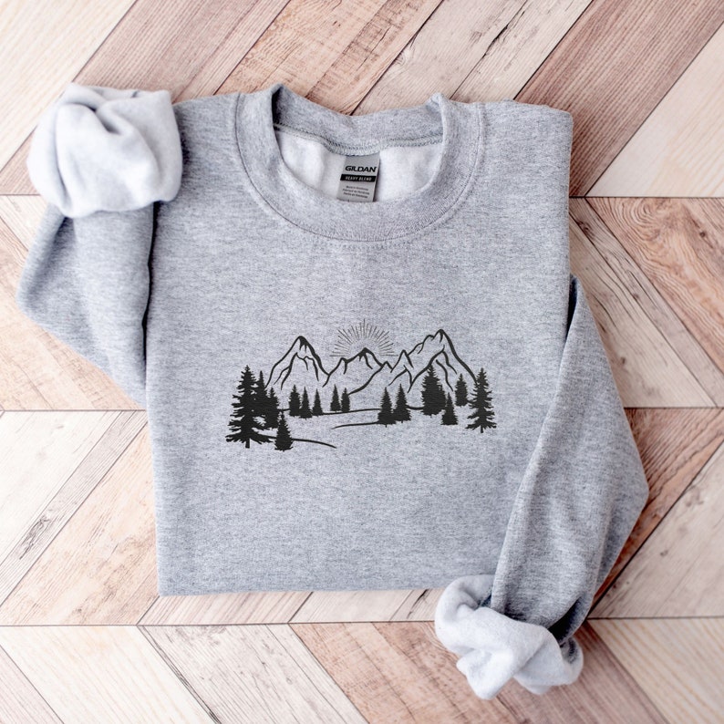 Op de afbeelding: Een grijs sweatshirt met een zwarte print van een bergketen en bomen. De afbeelding toont een zon die opkomt achter de bergen.